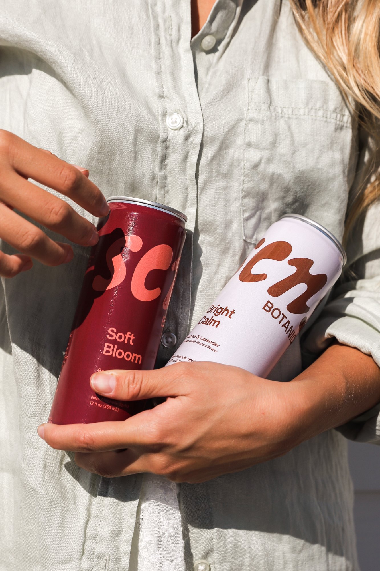 Two Sens Botanicals cans, Soft Bloom and Bright Calm, held against a linen shirt in soft sunlight